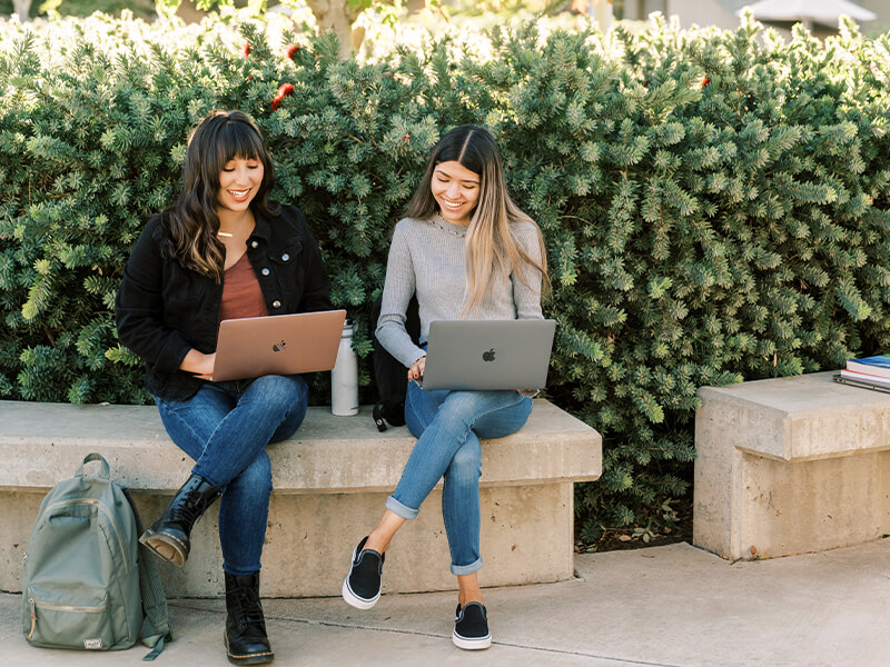 Two female students sitting outside while using their laptops