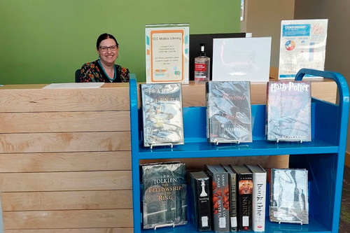 Megan the librarian sits behind a desk with a blue cart full of books in front of her.